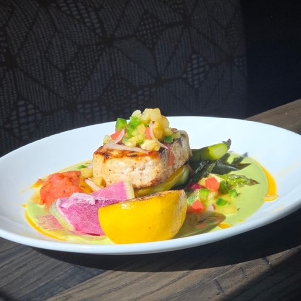 Grilled fish fillet served with green sauce, roasted vegetables, citrus wedges, and colorful pickled garnishes on a white plate.