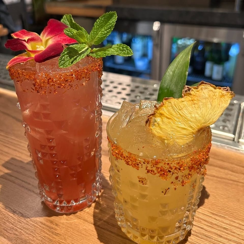 Two colorful cocktails with chili-salt rims, fresh mint, tropical flowers, pineapple garnish, and ice sit on a wooden bar.